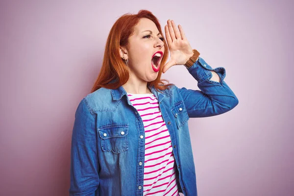 Beautiful redhead woman wearing denim shirt and striped t-shirt over isolated pink background shouting and screaming loud to side with hand on mouth. Communication concept.