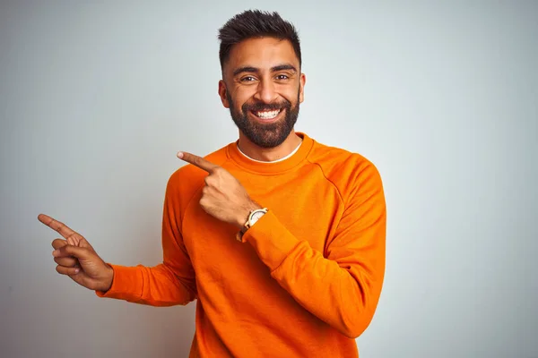 Young indian man wearing orange sweater over isolated white background smiling and looking at the camera pointing with two hands and fingers to the side.