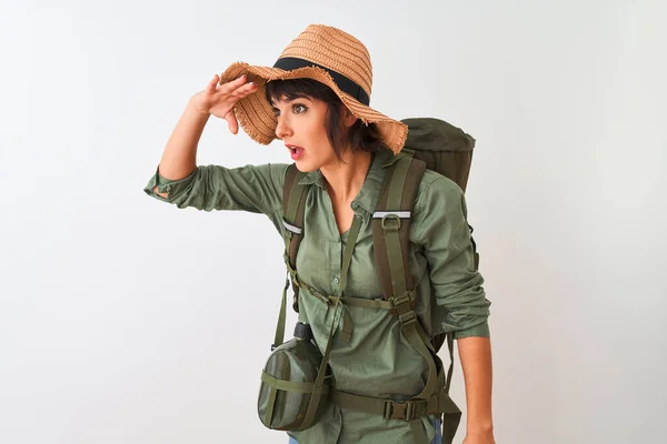 Hiker woman wearing backpack hat and water canteen over isolated white background very happy and smiling looking far away with hand over head. Searching concept.