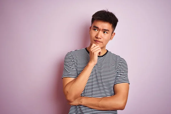 縞模様のTシャツを着た若いアジア系中国人男性がピンクの背景の上に立っていて あごは質問 集中的な表現について考えています 思慮深い顔で微笑んだ 疑いの概念 — ストック写真