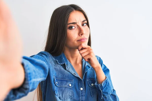 Beautiful woman wearing denim shirt make selfie by camera over isolated white background looking confident at the camera smiling with crossed arms and hand raised on chin. Thinking positive.