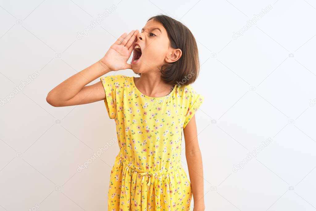 Young beautiful child girl wearing yellow floral dress standing over isolated white background shouting and screaming loud to side with hand on mouth. Communication concept.