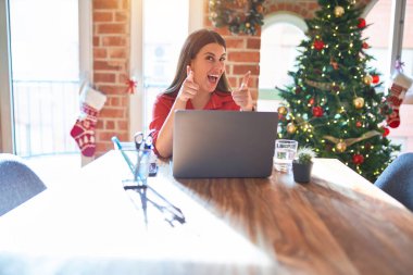 Güzel bir kadın evde dizüstü bilgisayarla oturup Noel ağacının etrafında mutlu ve komik yüzüyle kamerayı işaret ediyor. İyi enerji ve titreşimler.