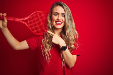 Genç sporcu raketle tenis oynuyor ve kırmızı izole arka planda bir top var. Elle ve parmakla göstererek çok mutlu.