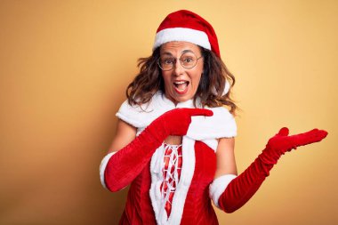 Orta yaşlı, güzel bir kadın, izole edilmiş sarı arka planda Noel Baba kostümü giyiyor. Kameraya hayret ediyor ve gülümsüyor..