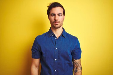 Yüzünde ciddi bir ifadeyle izole edilmiş sarı arka planda duran, sıradan tişörtlü, dövmeli genç yakışıklı bir adam. Basit ve doğal bir şekilde kameraya bakıyorsun..