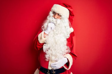 Orta yaşlı, yakışıklı, Noel Baba kostümü giymiş, izole edilmiş kırmızı arka planda duran bir adam bir soru hakkında endişeli ve gergin bir şekilde düşünüyor.