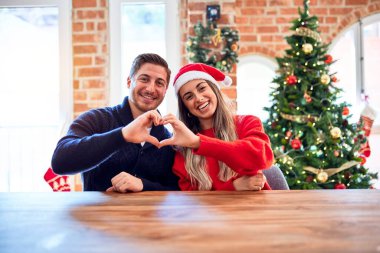 Noel Baba şapkası takan genç bir çift evdeki Noel ağacının etrafındaki sandalyeye ve masaya oturup gülümsüyor ve elleriyle kalp sembolü yapıyor. Romantik konsept.