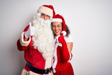 Orta yaşlı çift, Noel Baba kostümü giyip izole edilmiş beyaz arka planda sarılıyorlar. Elleriyle mutlu bir jest yapıyorlar. Kameraya bakıp başarıyı gösteren ifade onaylanıyor.