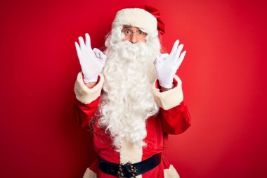 Noel Baba kostümü giymiş orta yaşlı, yakışıklı bir adam kırmızı arka planda dikiliyor. Şaşırmış ve şaşırmış görünüyor. Tamam sembolünü parmaklarıyla yapıyor. Çılgın ifade
