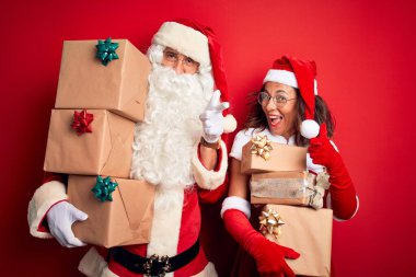 Noel Baba kostümü giyen orta yaşlı çift izole edilmiş kırmızı arka plan üzerinde hediye kulesi tutarak mutlu ve komik yüzlü parmaklarını kameraya doğrultuyor. İyi enerji ve titreşimler.