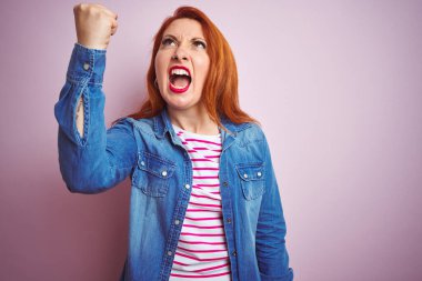 Güzel kızıl saçlı kadın kot gömlek ve çizgili tişört giyerek izole pembe arka planda kızgın kızgın ve öfkeli bir şekilde yumruk sallarken öfkeyle bağırıyor. Öfke ve agresif kavram.