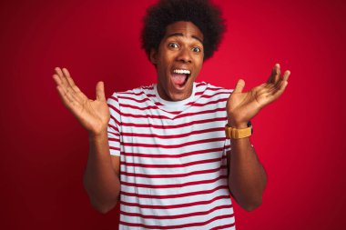 Afro saçlı, siyah tişörtlü genç Afro Amerikalı adam izole edilmiş kırmızı arka planda çılgınca kutlama yapıyor ve başarıyı kollarını kaldırıp heyecanla haykırarak haykırıyor. Kazanan konsept
