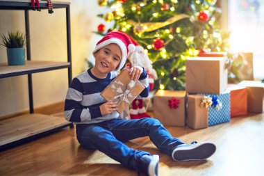 Sevimli çocuk mutlu ve kendinden emin gülümsüyor. Yerde oturmuş Noel Baba şapkası takıyor ve yüzünde gülümseme var. Evde Noel ağacının etrafında hediyeler tutuyor.