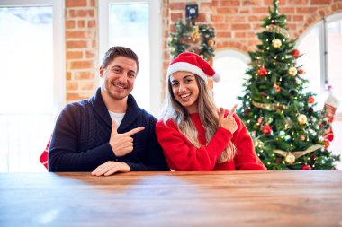 Noel Baba şapkası takan genç bir çift evdeki Noel ağacının etrafında oturmuş mutlu ve doğal bir ifadeyle elleri ve parmaklarıyla gülümseyerek Noel Baba şapkası takıyor.