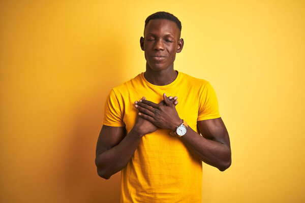 Young african american man wearing casual t-shirt standing over isolated yellow background smiling with hands on chest with closed eyes and grateful gesture on face. Health concept.