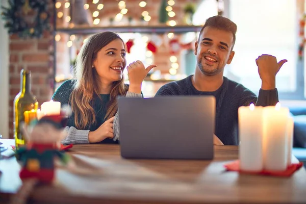 这对年轻漂亮的夫妇坐在家里用笔记本电脑装饰圣诞装饰品 面带微笑 面带微笑 手指头指向旁边 — 图库照片