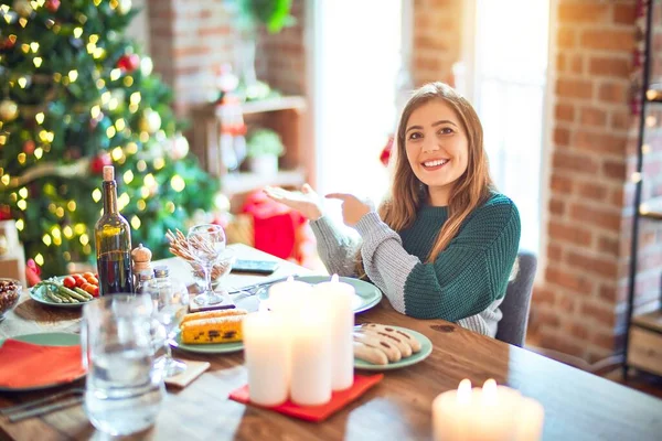 若いです美しい女性座って食べ物クリスマスツリーの周りに家に驚いて 手で提示しながら カメラに笑みを浮かべて指で指して — ストック写真