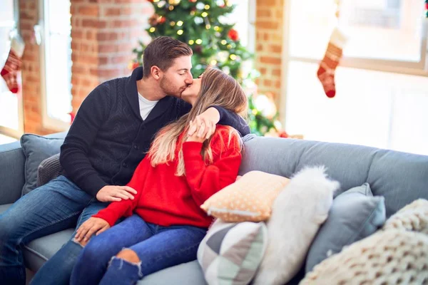 年轻漂亮的夫妇面带微笑 既快乐又自信 坐在沙发上拥抱和亲吻着家里的圣诞树 — 图库照片