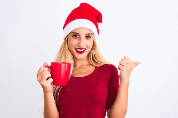 漂亮的女人戴着圣诞圣诞圣诞礼帽 站在孤独的白色背景上喝咖啡 手指着 带着大拇指站在一旁 笑容满面 — 图库照片
