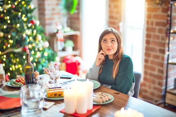 若い美しい女性の質問について考える顎の手で自宅でクリスマスツリーの周りの食品を食べて座って 集中的な表現 思慮深い顔で微笑んだ 疑いの概念 — ストック写真