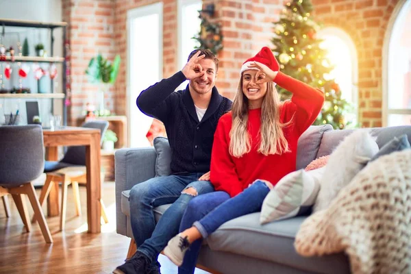 若いですカップル身に着けていますサンタクラスの帽子座っていますザソファ周りにクリスマスツリーでホーム行うOkジェスチャーとともに手笑顔 目を通して指で見て幸せな顔 — ストック写真