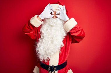 Orta yaşlı yakışıklı bir adam, Noel Baba kostümü giymiş kırmızı arka planda dikiliyor. Dürbün gibi güzel hareketler yapıyor, gözleri parmaklarından bakıyor. Çılgın ifade.