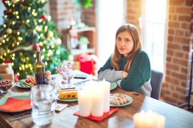 Young beautiful woman sitting eating food around christmas tree at home skeptic and nervous, disapproving expression on face with crossed arms. Negative person. clipart