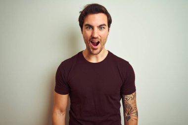 İzole edilmiş beyaz arka plan üzerinde mor tişört giyen yakışıklı genç bir adam korku, korku ve heyecan dolu bir yüz ifadesiyle korkmuş ve şok olmuştu..
