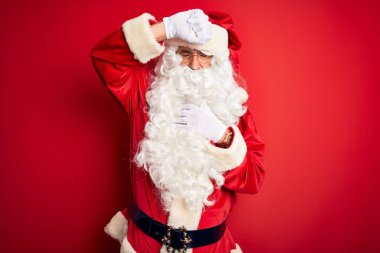 Noel Baba kostümü giyen orta yaşlı yakışıklı adam izole edilmiş kırmızı arka planda duruyor. Hastalık, ateş, grip, soğuk algınlığı ve virüs için alnına dokunuyor.