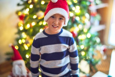 Sevimli çocuk mutlu ve kendinden emin gülümsüyor. Noel Baba şapkası takmış, Noel ağacının etrafında gülümsüyor ve evde bir sürü hediye var.