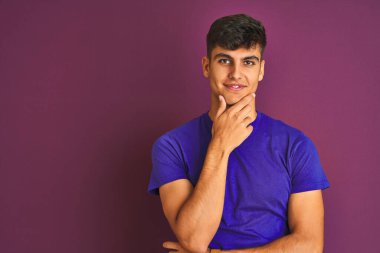 İzole edilmiş mor arka planda duran tişört giyen genç Hintli adam kameraya kollarını kavuşturup gülümseyerek ve çenesini kaldırarak bakıyor. Olumlu düşünüyorum..