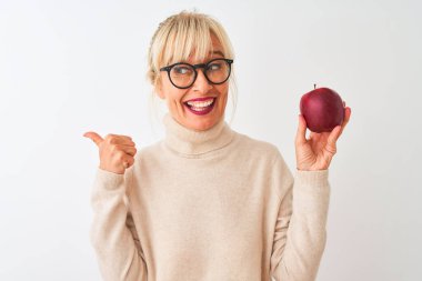 Elma tutan orta yaşlı bir kadın soyutlanmış beyaz arka plan üzerinde duruyor ve gülümseyen bir gülümsemeyle baş parmağıyla işaret ediyor.