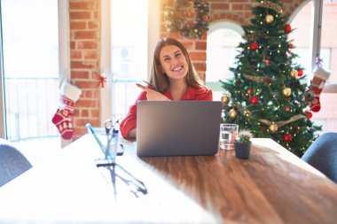 Güzel bir kadın evinde dizüstü bilgisayarla oturuyor Noel ağacının etrafında neşeli bir gülümsemeyle elleri ve parmaklarıyla mutlu ve doğal bir ifadeyle yüzünü gösteriyor.