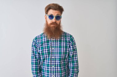İzole edilmiş beyaz arka plan üzerine gündelik gömlek ve güneş gözlüğü takan genç kızıl saçlı adam yüzünde ciddi bir ifadeyle rahatlamış. Basit ve doğal bir şekilde kameraya bakıyorsun..