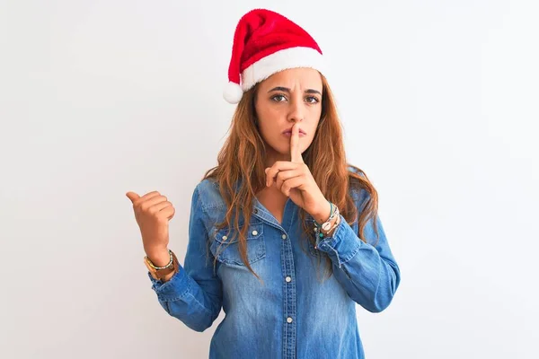 孤立した背景の上にクリスマスの帽子を身に着けている若い美しい赤毛の女性は 唇に手で側に指して指で静かになるように求めています 沈黙と秘密の概念 — ストック写真