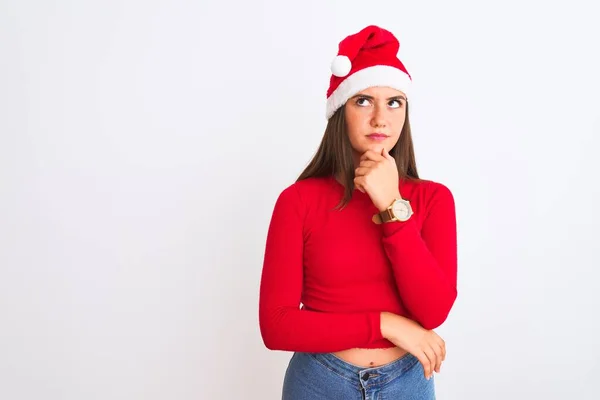 若い美しい女の子の質問について考えてあごの手で隔離された白い背景の上に立ってクリスマスサンタの帽子をかぶって 包括的な表現 思慮深い顔で微笑んだ 疑いの概念 — ストック写真
