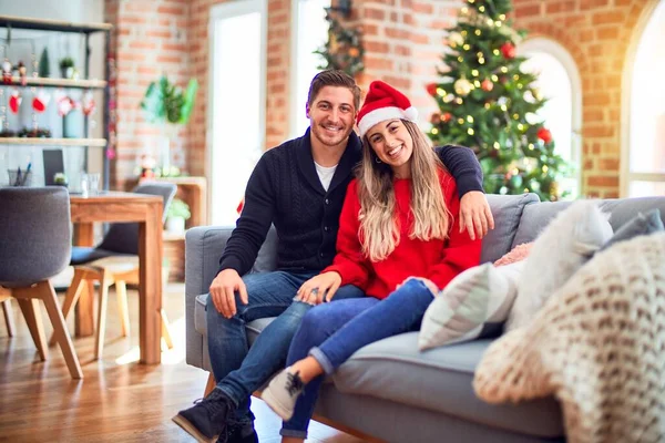 一对年轻的夫妇 头戴圣爪帽 坐在沙发上 围着圣诞树 脸上挂着快乐而凉爽的笑容 幸运的人 — 图库照片