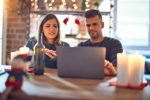 Young beautiful couple sitting using laptop around christmas decoration at home disgusted expression, displeased and fearful doing disgust face because aversion reaction. With hands raised. Annoying concept.