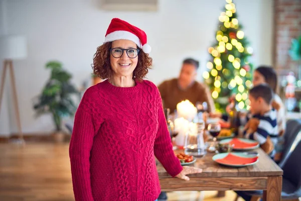中年の美しい女性が幸せと自信を笑顔 自宅で家族と一緒にクリスマスを祝うサンタクラスの帽子を身に着けてポーズ — ストック写真