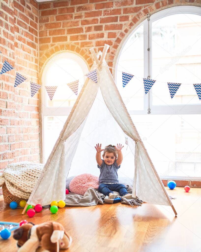 Beautiful toddler sitting on the floor inside tipi with hands raised smiling at kindergarten