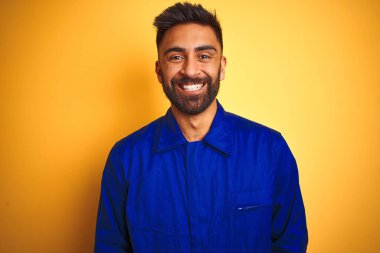 Yüzünde mutlu ve havalı bir gülümsemeyle izole edilmiş sarı arka plan üzerinde üniforma giyen yakışıklı Hintli işçi. Şanslı kişi..