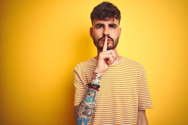 Dövmeli, çizgili tişörtlü genç adam izole edilmiş sarı arka planda duruyor ve sessiz olmak istiyor. Sessizlik ve gizli konsept.