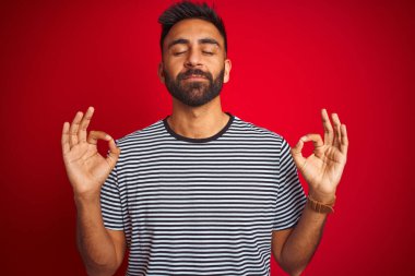 İzole edilmiş kırmızı arka plan üzerinde denizci çizgili tişört giyen genç ve yakışıklı Hintli adam rahatla ve gözleri kapalı parmaklarıyla meditasyon hareketi yaparken gülümse. Yoga konsepti.