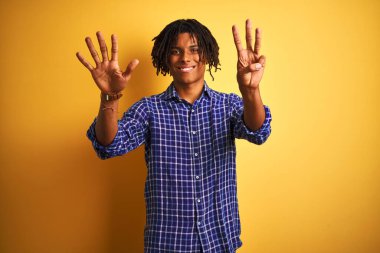 Rastgele tişörtlü Afro bir adam izole edilmiş sarı arka planda dikiliyor ve 8 numaralı parmaklarıyla işaret ederken kendinden emin ve mutlu gülümsüyor..