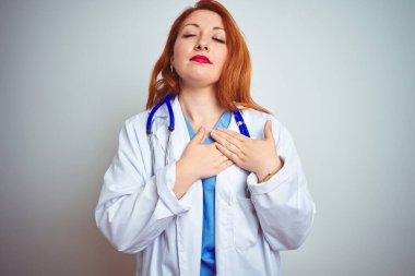 Young redhead doctor woman using stethoscope over white isolated background smiling with hands on chest with closed eyes and grateful gesture on face. Health concept.