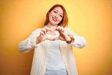İzole edilmiş sarı arka planda duran çizgili tişört giyen güzel kızıl saçlı kadın. Aşkla gülümsüyor. Elleriyle kalp sembolü ve şekli gösteriyor. Romantik konsept.