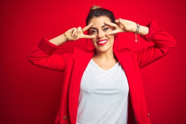 Genç ve güzel bir iş kadını kırmızı izole edilmiş arka planda duruyor. Yüzünde parmaklarıyla barış sembolü yapıyor. Gülümsüyor, zaferini gösteriyor.