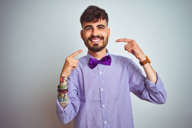 Mor gömlekli ve papyonlu genç adam izole edilmiş beyaz arka planda gülümseyen ve parmaklarıyla dişlerini ve ağzını gösteren. Diş sağlığı konsepti.