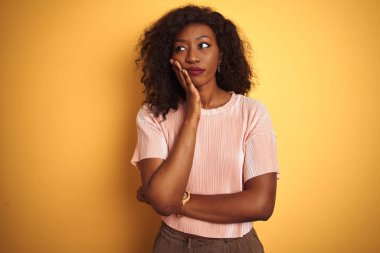 Pembe tişört giyen Afro-Amerikalı kadın izole edilmiş sarı arka planda duruyor. Kollarını çapraz tutarak depresyondan sıkılmış ve yorgun görünüyor..
